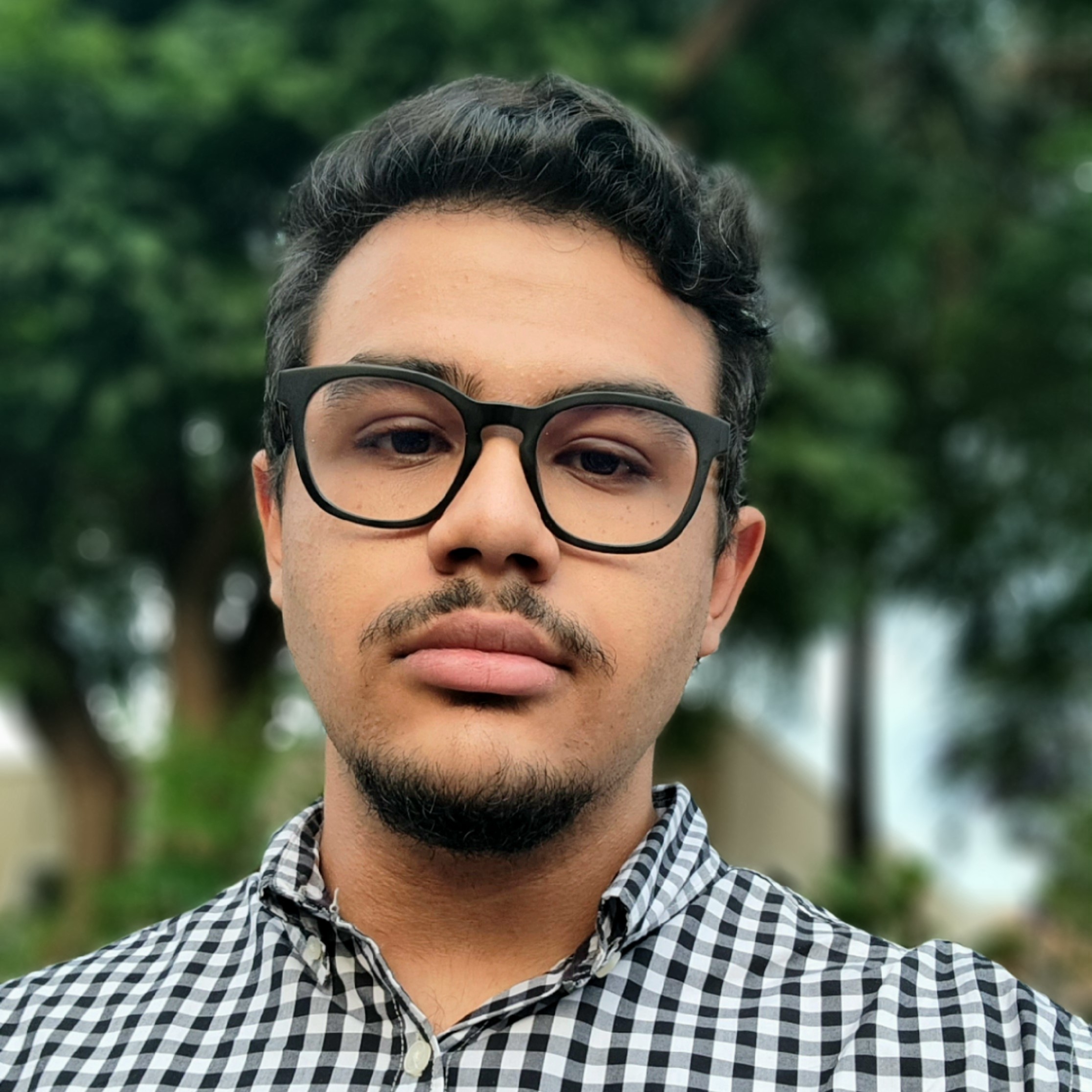 Minha selfie, no fundo tem algumas arvores, eu estou com um olhar sério, usando um óculos redondo e preto